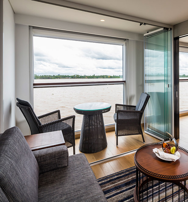 A Deluxe Suite with a queen bed and an elegant lounge area, framed by floor-to-ceiling windows with views of the passing river.