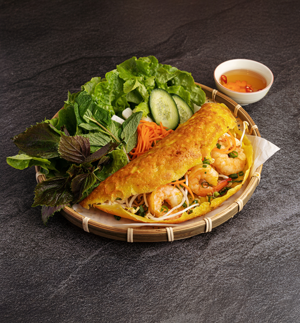 Vietnamese bánh xèo savoury pancake with shrimp, bean sprouts, fresh herbs, and dipping sauce on a bamboo plate.